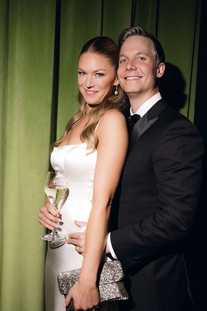 Bride and groom posing close together in front of rich emerald green velvet curtains, with the bride holding a wine glass and wearing opera gloves.