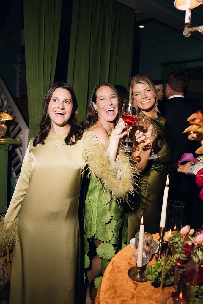 Three women in elegant dresses smiling and holding drinks inside a warmly decorated room with green drapes and candlelit tables at Bradford House.