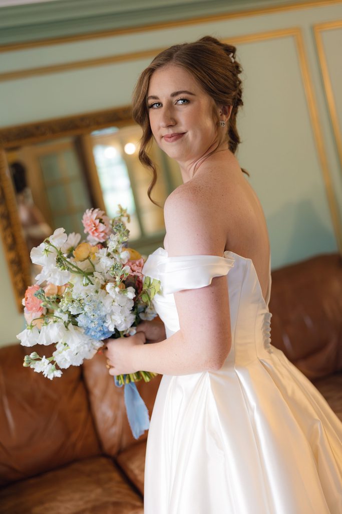 Bride in an off-the-shoulder gown holding a pastel bouquet with blush, cream, and soft blue flowers, posing indoors at Dresser Mansion.