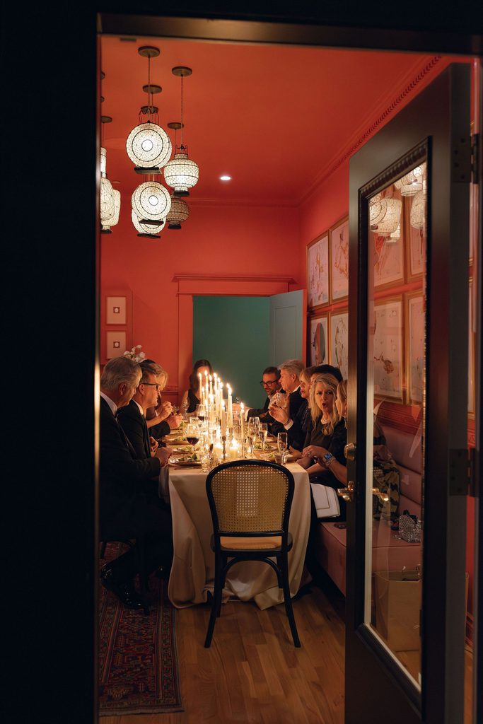 Warmly lit wedding reception setup inside a coral-pink room at Bradford House, featuring lit taper candles along a dining table, framed artwork, and globular pendant light fixtures overhead.