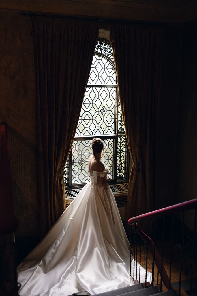 Full-length silhouette portrait of the bride in her ballgown standing at a tall leaded arched window frame flanked by dark curtains inside Dresser Mansion.