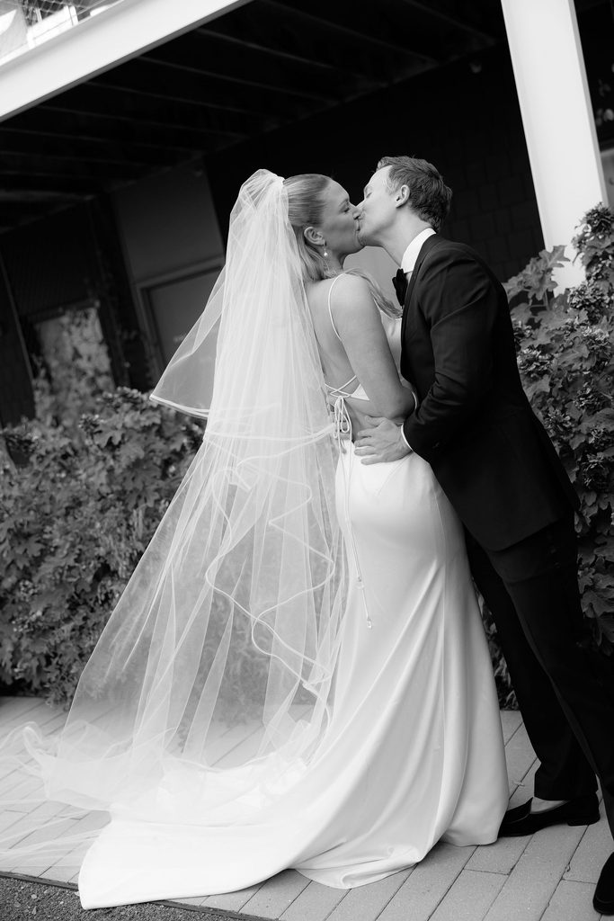 Black-and-white portrait of the bride and groom kissing outdoors on a porch at Bradford House, with the bride wearing a long white veil.