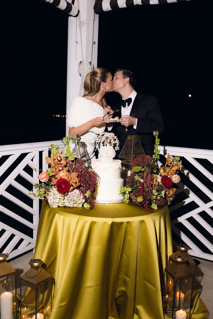 Bride and groom kissing outdoors at night behind a floral-adorned wedding cake on a green tablecloth under a black-and-white striped awning.