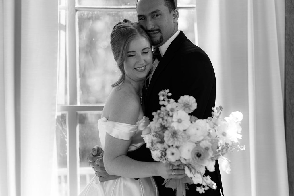 Black-and-white portrait of the bride holding a pastel floral bouquet and smiling while leaning gently against the groom in his black tuxedo.