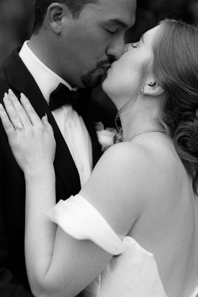 Close-up black-and-white photograph of the bride and groom kissing, highlighting the bride's engagement ring and drop earrings.