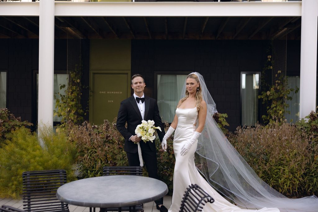 Bride and groom standing together outside near stone steps and trees, with the bride in a strapless gown with opera gloves holding a white bouquet, and the groom wearing a black tuxedo.