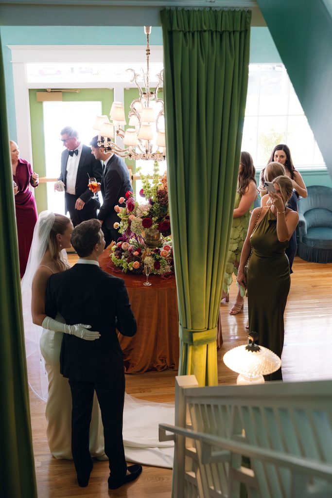 Bride and groom viewed through an open doorway inside Bradford House, framed by green curtains with guests mingling in the brightly decorated interior.